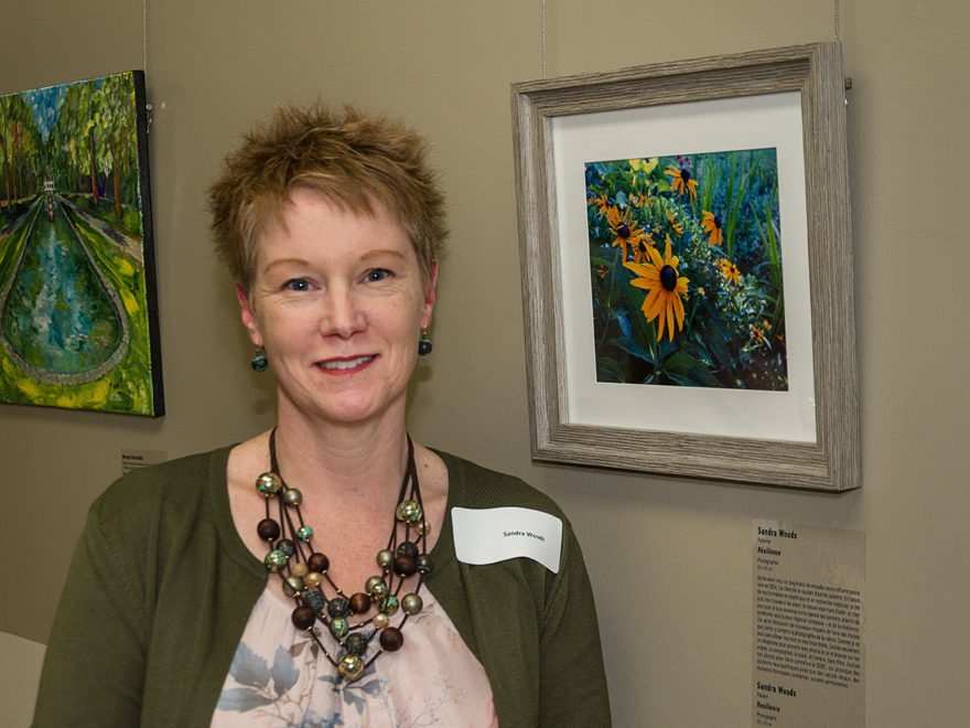 a woman standing beside a framed image of flowers, in an art-gallery style setting. A name tag on her shoulder says: Sandra Woods