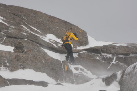 a man snowshoeing along a narrow ledge of a rock face, with heavy wind blowing up the snow around him
