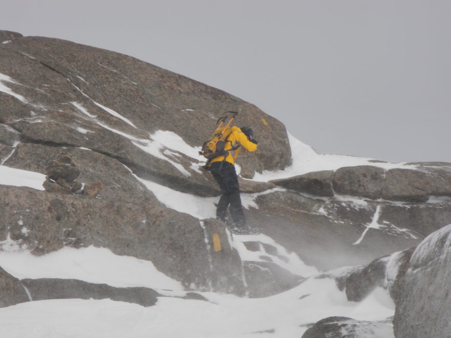 a man snowshoeing along a narrow ledge of a rock face, with heavy wind blowing up the snow around him