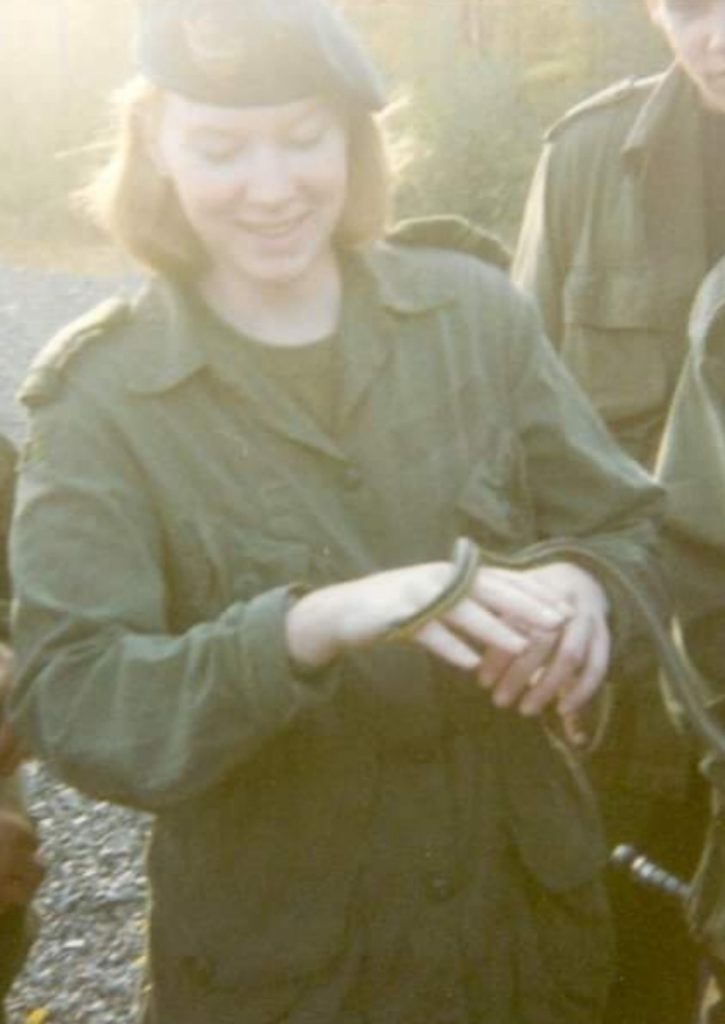 a female military officer wearing combat fatigues, standing with her hands at waist level, letting a small snake holding a small snake move between her two hands