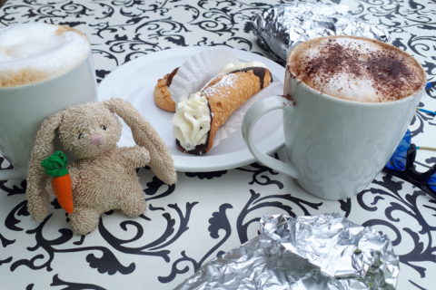 a small plush rabbit, on a table with two cups of cappuccino and some cookies.