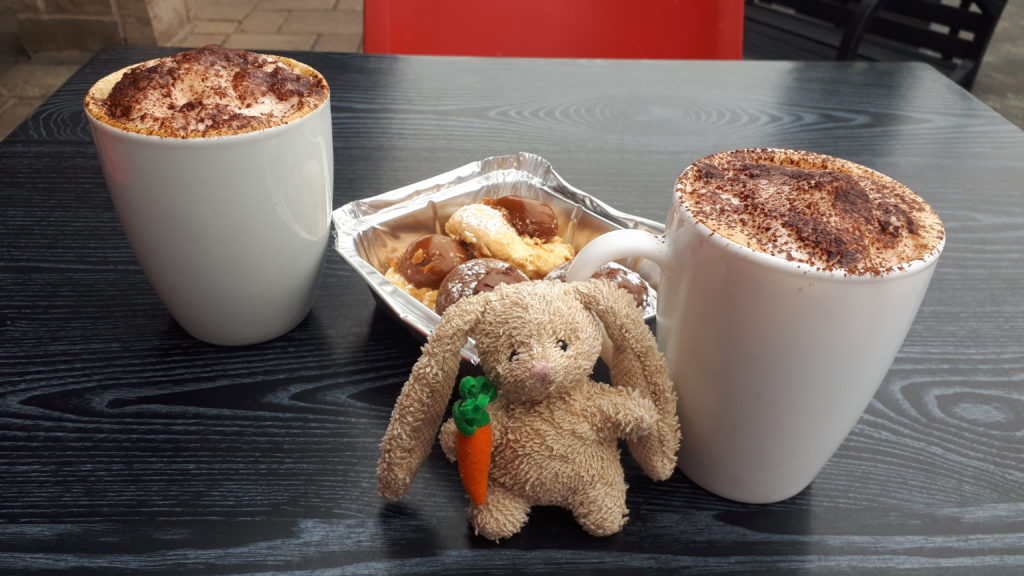 a small plush rabbit, on a table with two cups of cappuccino coffee and some cookies