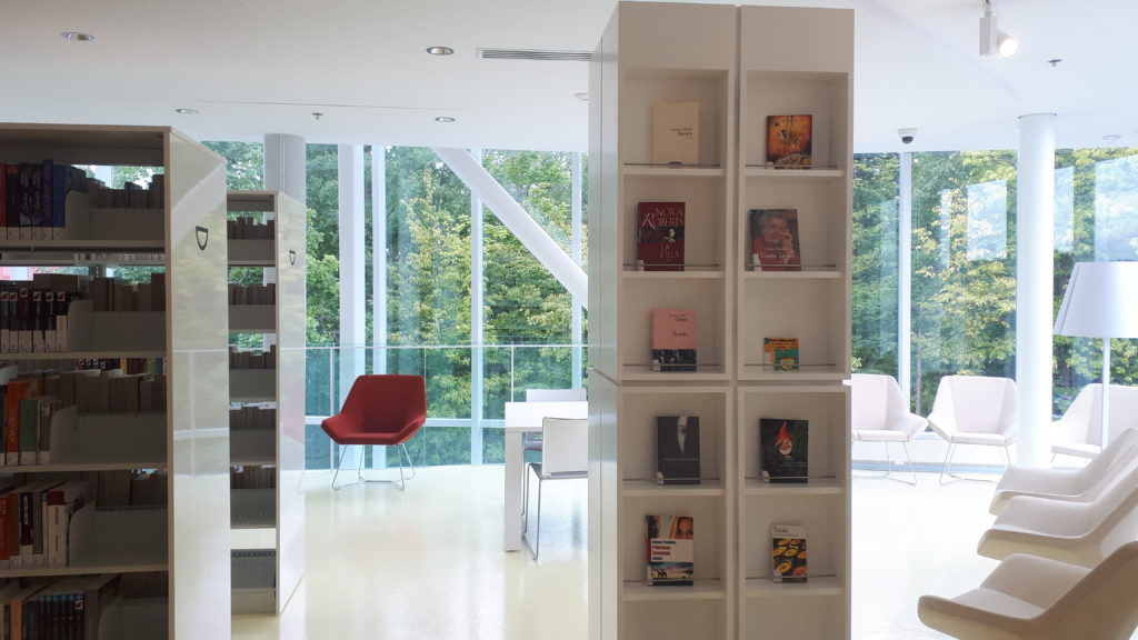 a seating area in a library, with some books showcased on museum-style shelves, There are 10 comfortable chairs along a window, with another 4 seats around a small table.