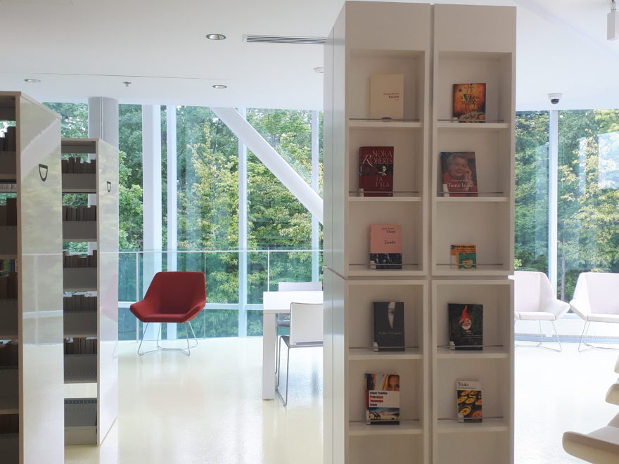 a seating area in a library, with some books showcased on museum-style shelves, There are 10 comfortable chairs along a window, with another 4 seats around a small table.