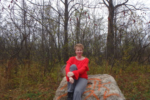 the author, sitting on a rock in a forest and wearing a bright orange sweater for CRPS awareness day