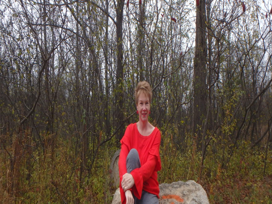 the author, sitting on a rock in a forest and wearing a bright orange sweater for CRPS awareness day
