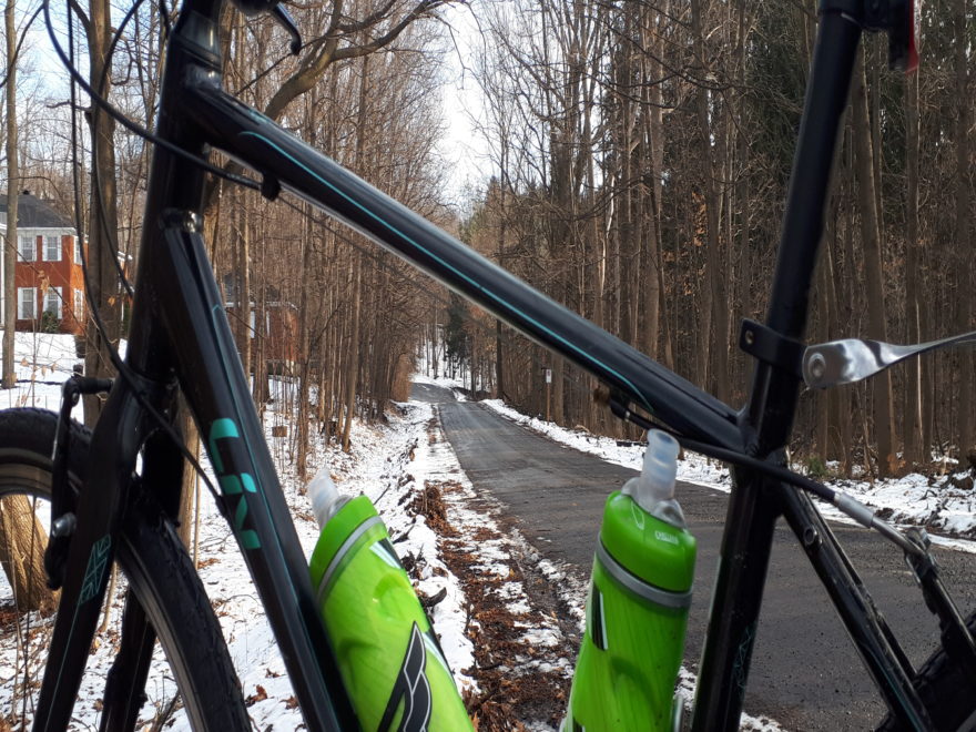 a woman's bicycle, at the bottom of a small hill in a forest. There is snow on the ground, but not on the road which is wet.