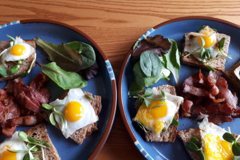 two plates of sunny-side up quail eggs on French bread, with sunflower sprouts, mixed baby lettuce leaves, and bacon