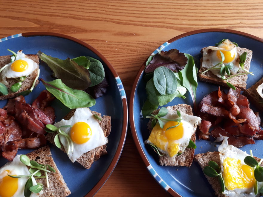 two plates of sunny-side up quail eggs on French bread, with sunflower sprouts, mixed baby lettuce leaves, and bacon