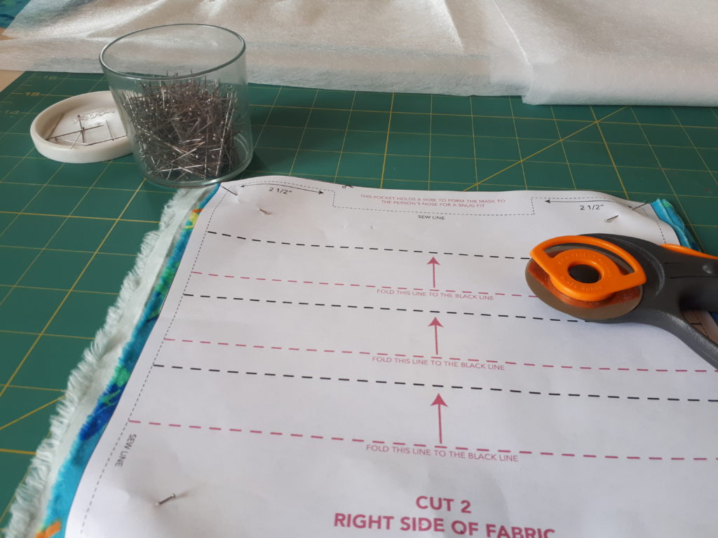 a piece of fabric on a cutting board, with a paper sewing pattern for a surgical-style face mask pinned on top of it. There are a container of sewing pins and a cutting tool in the background