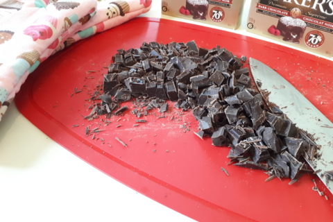 an entire package of Baker's chocolate, chopped into small pieces on a chopping mat. In the background is a set of oven mitts printed with images of cupcakes, along with two boxes of 70 percent dark chocolate. There's a large knife in the foreground