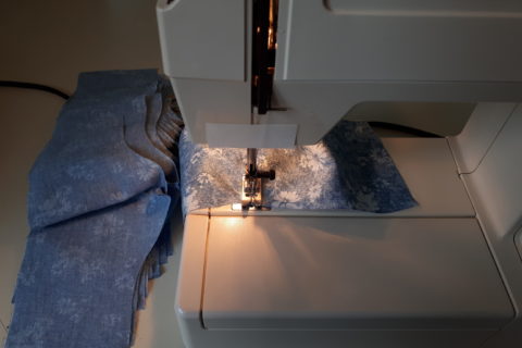 an old portable sewing machine, with a partially-completed cotton mask being sewn. Alongside it are a pile of ten or so other partially-sewn masks, awaiting their turn at the sewing machine