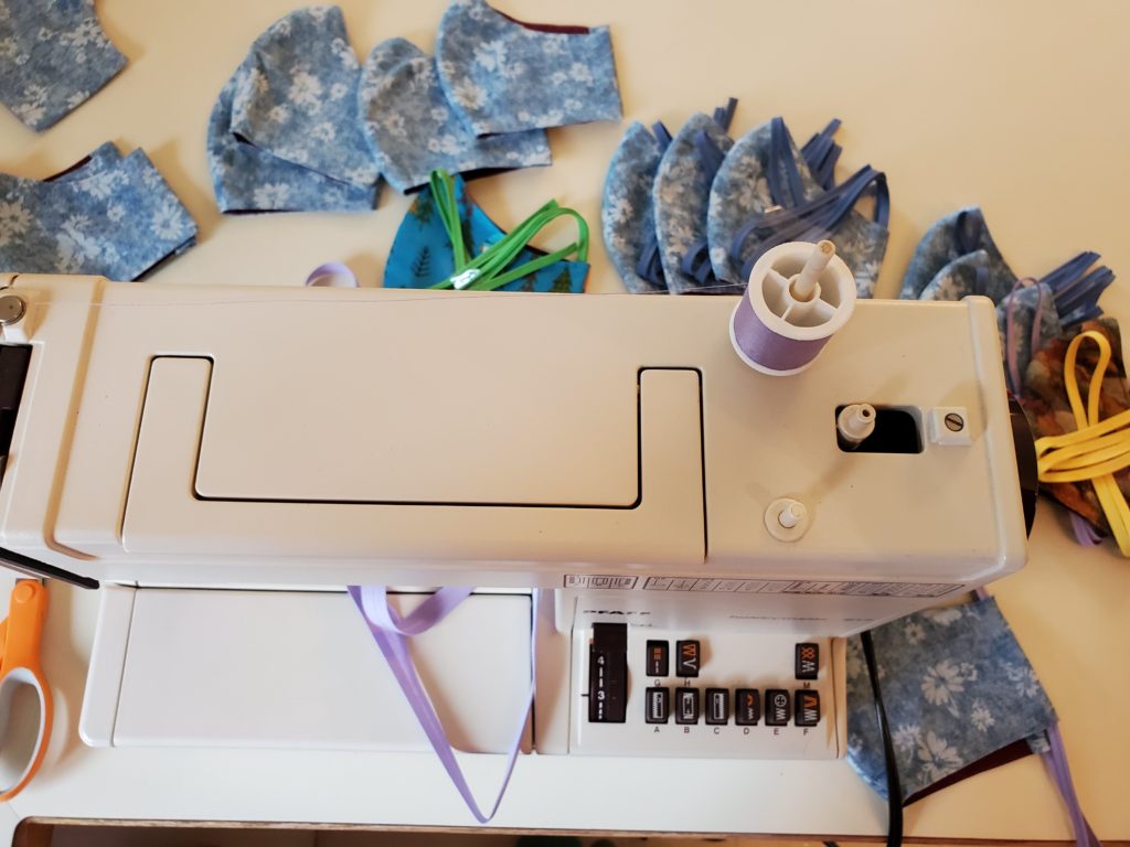 A portable sewing machine, surrounded by 16 partially-completed home-sewn cotton face masks