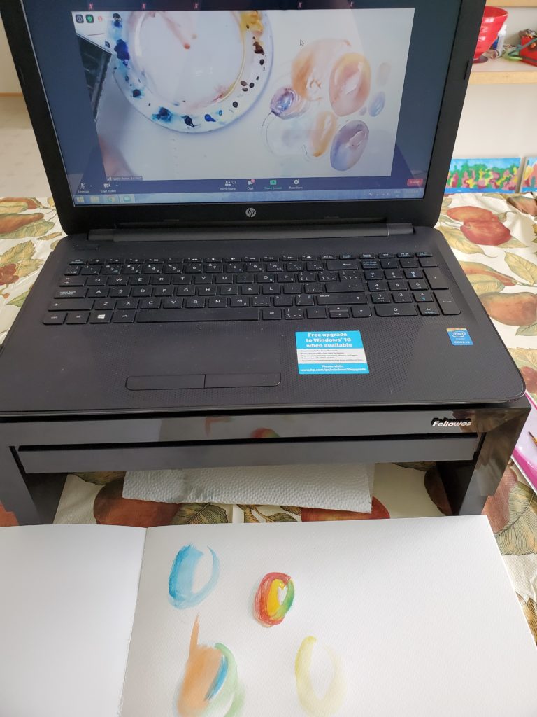 A laptop computer on a dining table, showing a watercolour art tutorial hosted by Art Safari in Engand. In front of the computer is a piece of watercolour paper on which the beginner has painted some designs.