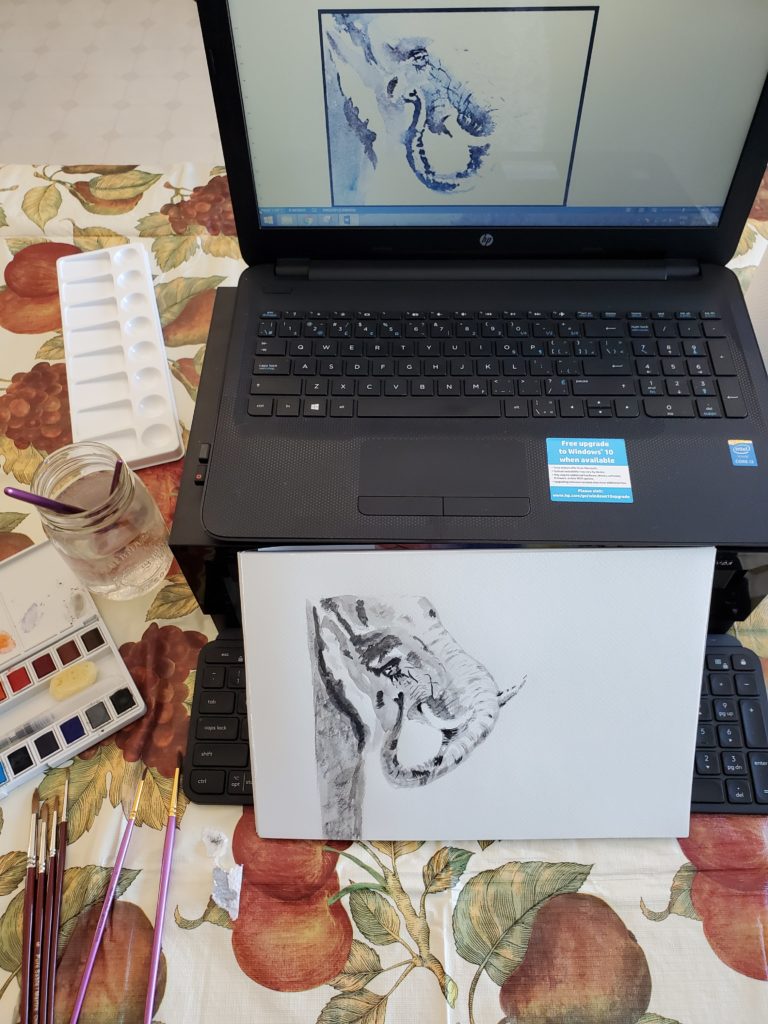 An amateur watercolour painting of an elephant, in front of a computer screen showing a similar image as a model. There are watercolour paints and brushes lying on the table, alongside the painting.