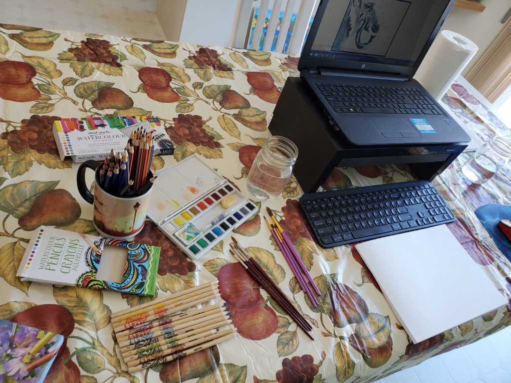 A dining room table covered with a waterproof cloth, with several sets of watercolour paints and brushes beside a blank pad of watercolour paper.. There is a laptop computer on the table, showing an image of an elephant on the screen.