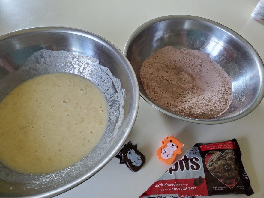 Two mixing bowls containing the dry and wet ingredients for homemade chocolate banana muffins, beside a package of dark chocolate chips