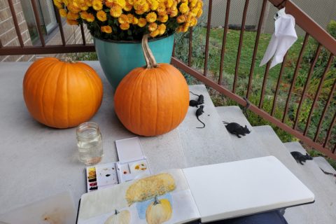 Two pumpkins and a large pot of fall flowers outside someone's front door, and a sketchbook into which these items are being painted in watercolours