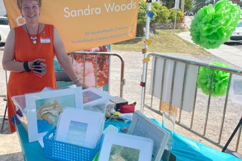 a woman standing behind a table covered in boxes of watercolour paintings and chronic pain awareness information, at the What the Pop! 2022 outdoor art show