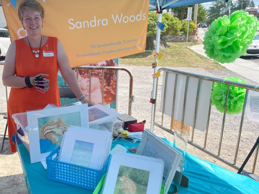 a woman standing behind a table covered in boxes of watercolour paintings and chronic pain awareness information, at the What the Pop! 2022 outdoor art show