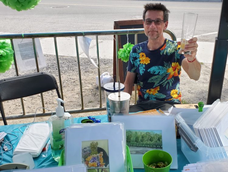 A man seated behind and artist's table at the What the Pop! outdoor art show, holding up his glass as a hello