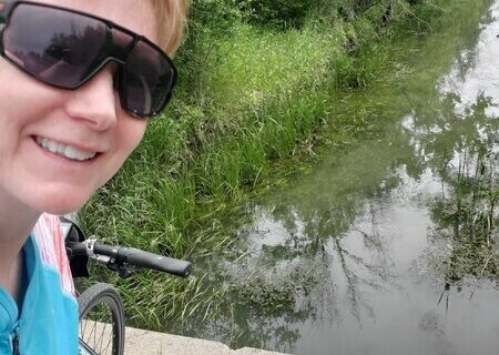 A woman standing beside a bicycle, in front of a creek. Balanced on the saddle of the bike is a painting of the creek.