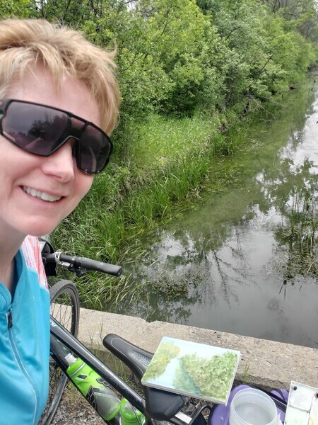 A woman standing beside a bicycle, in front of a creek. Balanced on the saddle of the bike is a painting of the creek.