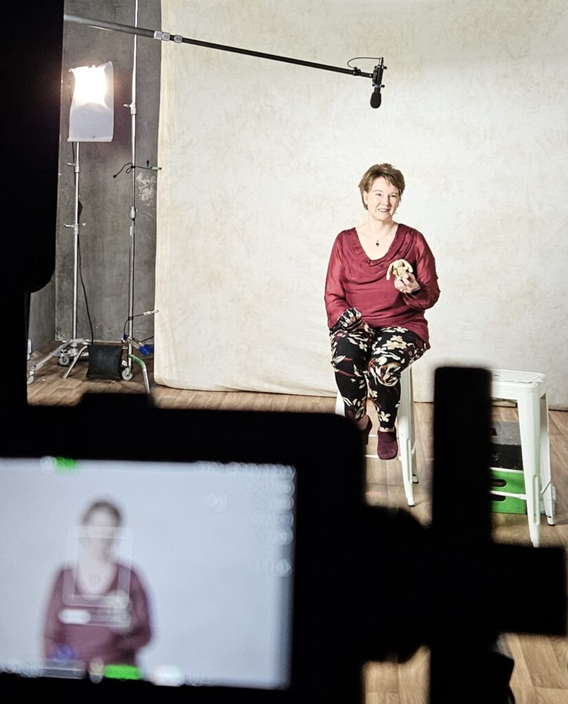 Sandra Woods seated on a stool on a film set with a blank screen behind her, on the set of You Can't Ask That